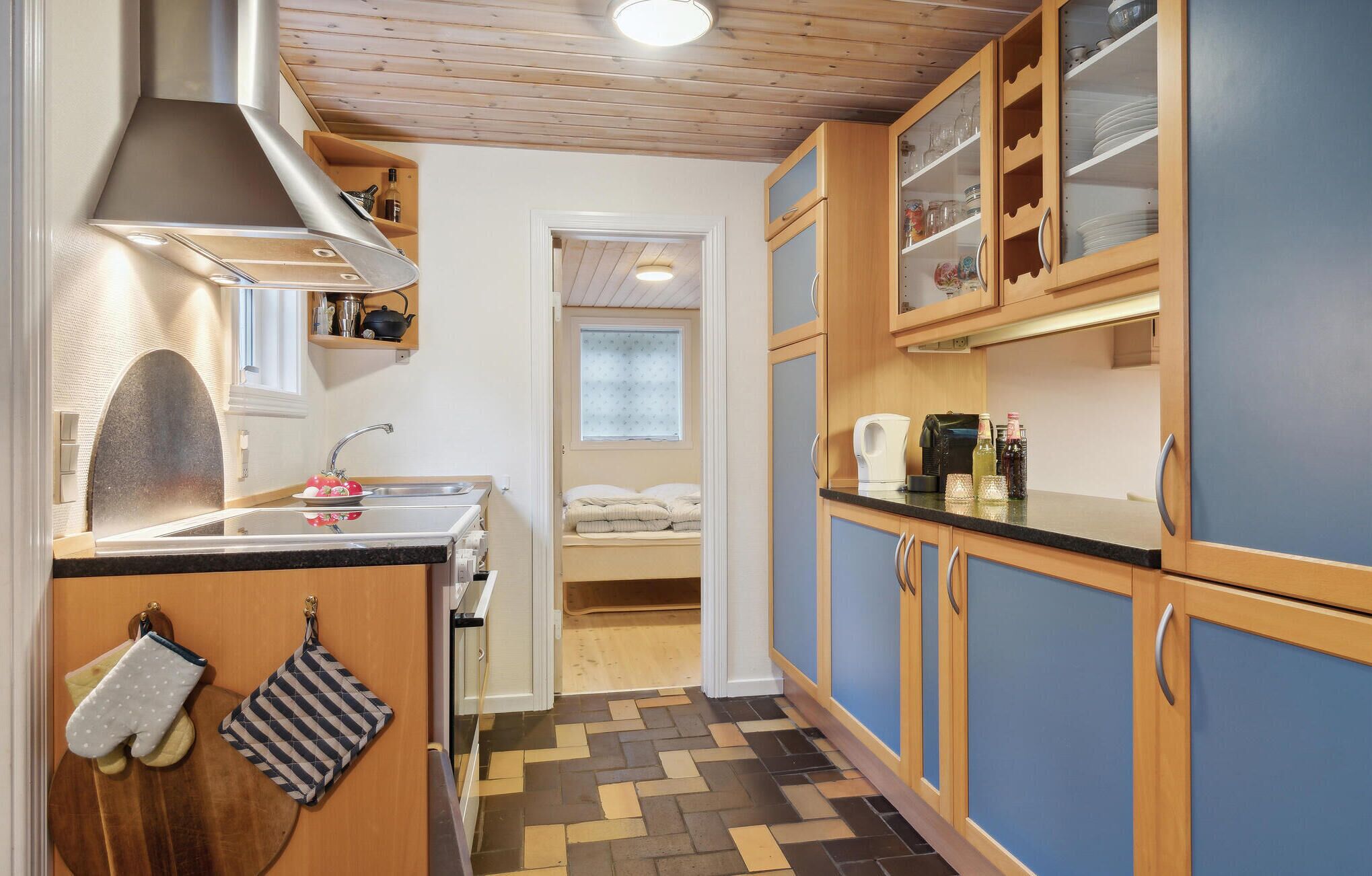 Una cucina funzionale con mobili in legno, un lavello e un fornello. Si vede una piccola finestra e un ingresso che conduce a una camera da letto.