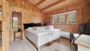 Et soveværelse i en træhytte med seng, sengebord, lampe og et vindue med udsigt til træer.