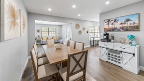 Uma sala de jantar espaçosa com uma mesa e cadeiras de madeira, um aparador branco com gavetas e um quadro grande com uma paisagem de praia.