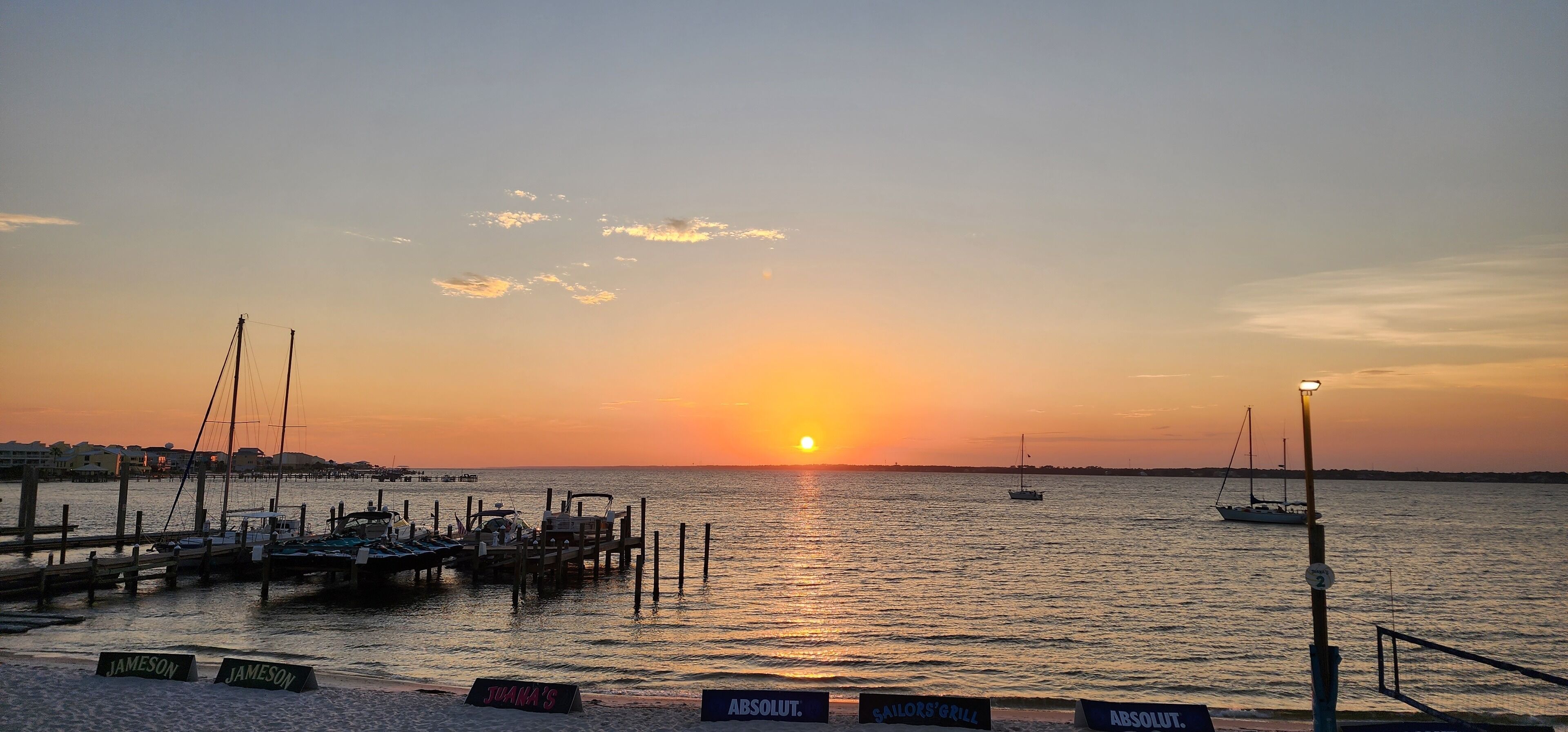 Un coucher de soleil sur une marina où sont amarrés des bateaux, avec une jetée et une enseigne indiquant « JAMA’S ».