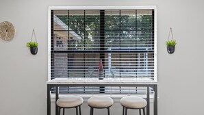 Uma cozinha com janela, uma mesa e três banquetas. Há duas plantas em vasos na parede.