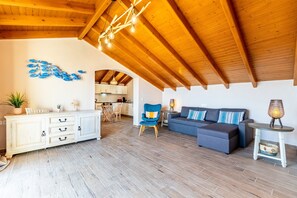 Amplia sala de estar con techo de madera alto, un gabinete blanco con cajones, un sillón azul, un sofá gris y una pequeña mesa con una lámpara.