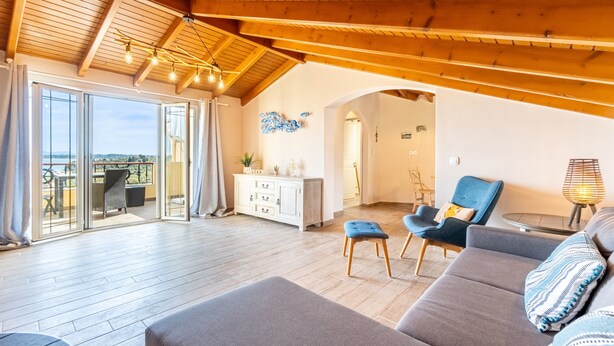 Een ruime woonkamer met een houten plafond, een bank, een blauwe fauteuil, een glazen deur naar een balkon en een kleine tafel met een lamp.