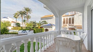 A balcony with white railings, a white table, and chairs, overlooking a garden with palm trees and a building with a blue roof. - Holiday Home 'Bungalow 213' with Shared Pool, Wi-Fi and Air Conditioning (Santa Pola)