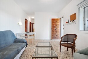 Un salon comprenant un canapé, une table basse en verre, un fauteuil en osier, une table blanche et une porte en bois.