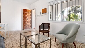 Une pièce avec une table en verre, une chaise en osier, un fauteuil à motifs, une porte en bois et une fenêtre avec des barreaux.