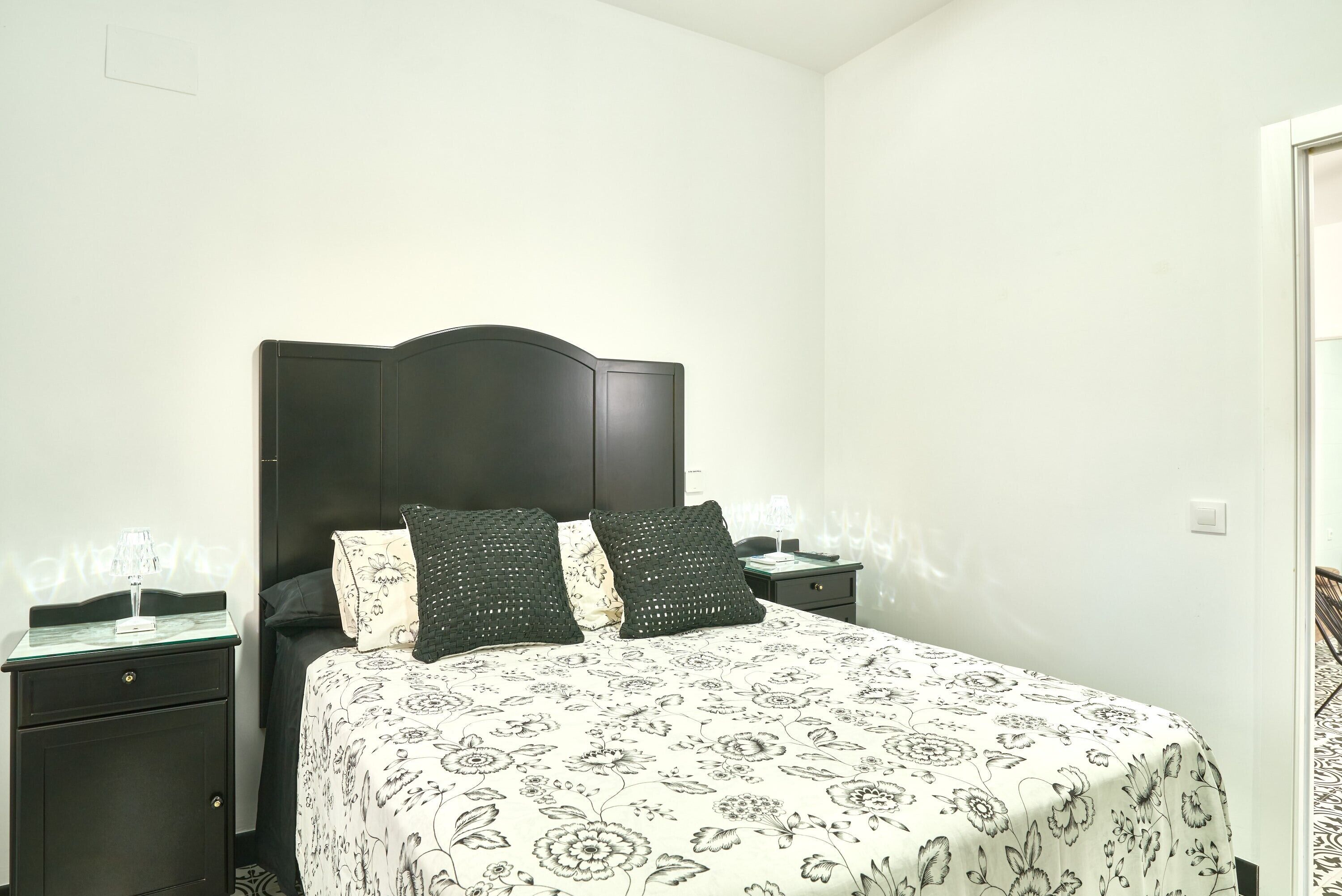 Ein Schlafzimmer mit einem Bett, zwei Nachttischen, einem dunklen Kopfteil und einer zudeckenden Bettwäsche mit Blumenmuster.