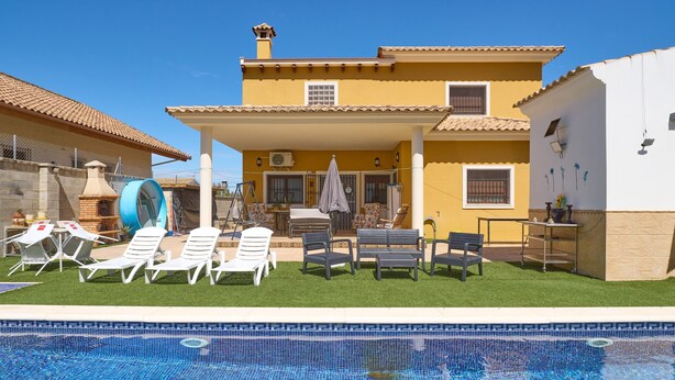 A poolside area with lounge chairs, a barbecue grill, and a house with a tiled roof. - Holiday Home 'Marisol' with Shared Pool, Wi-Fi & Air Conditioning (Puente Genil)