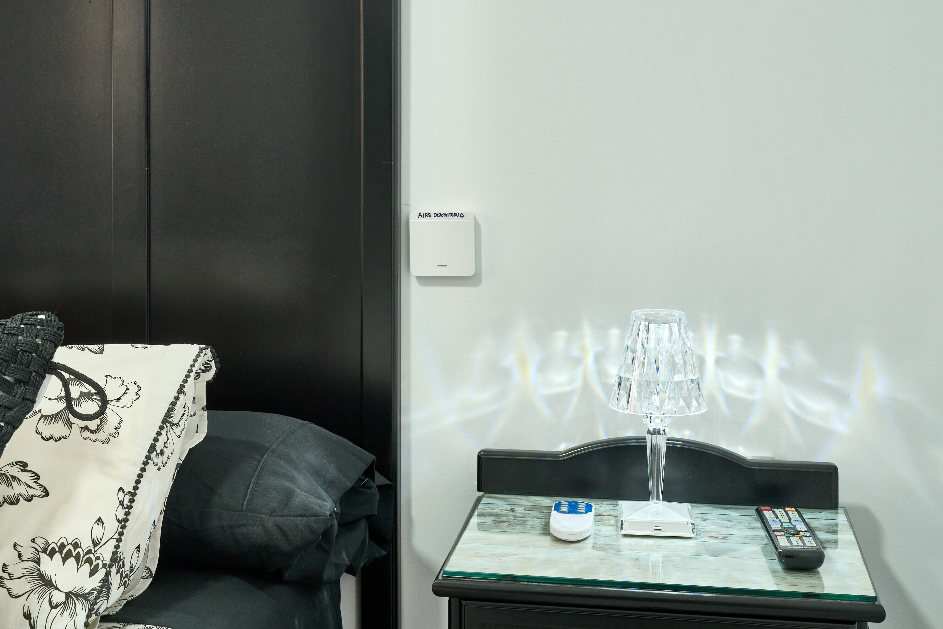A hotel room with a bed, a bedside table with a lamp, a remote control, and a thermostat on the wall.