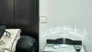 A hotel room with a bed, a bedside table with a lamp, a remote control, and a thermostat on the wall.