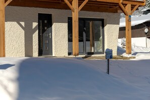 Um edifício com o chão coberto de neve, uma pérgula de madeira e uma porta com janela.