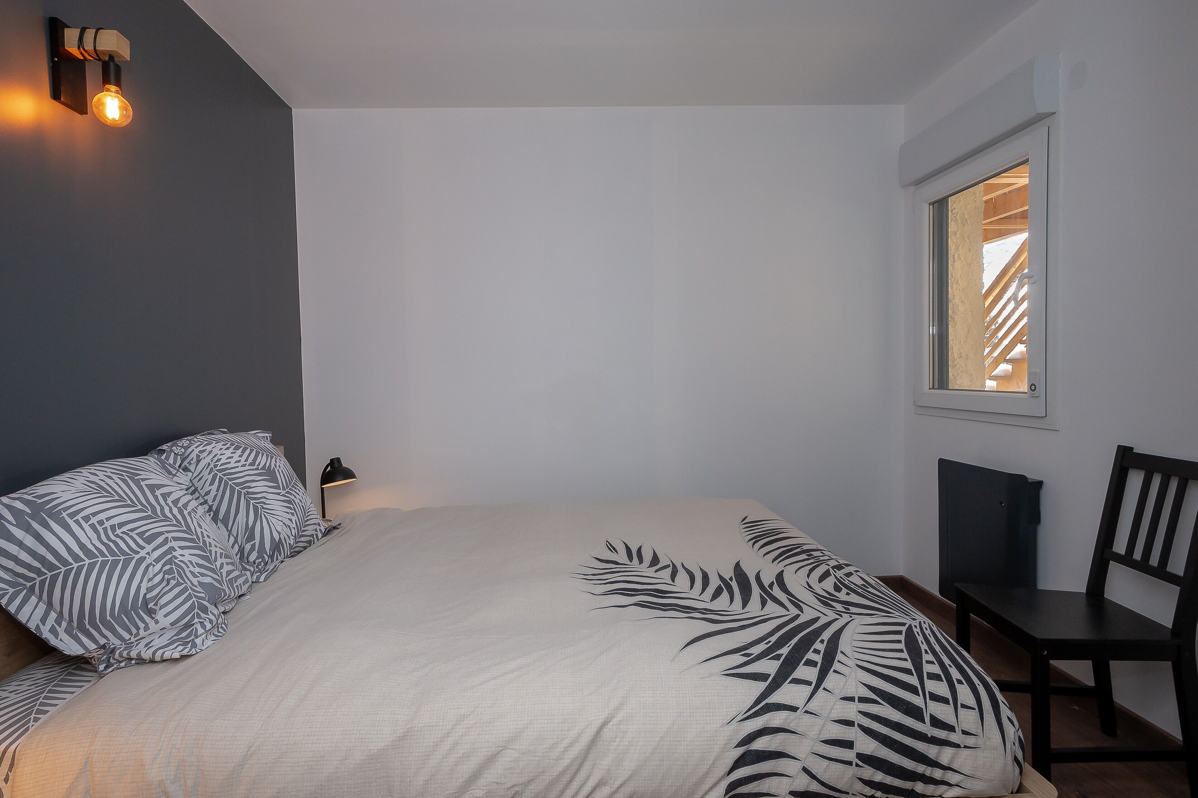 Une chambre à coucher comprenant un lit, une chaise, un bureau, une fenêtre et un luminaire fixé au mur.