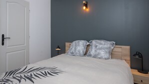 Un dormitorio con cama, dos almohadas, una mesita de noche, una lámpara de pared y una puerta.