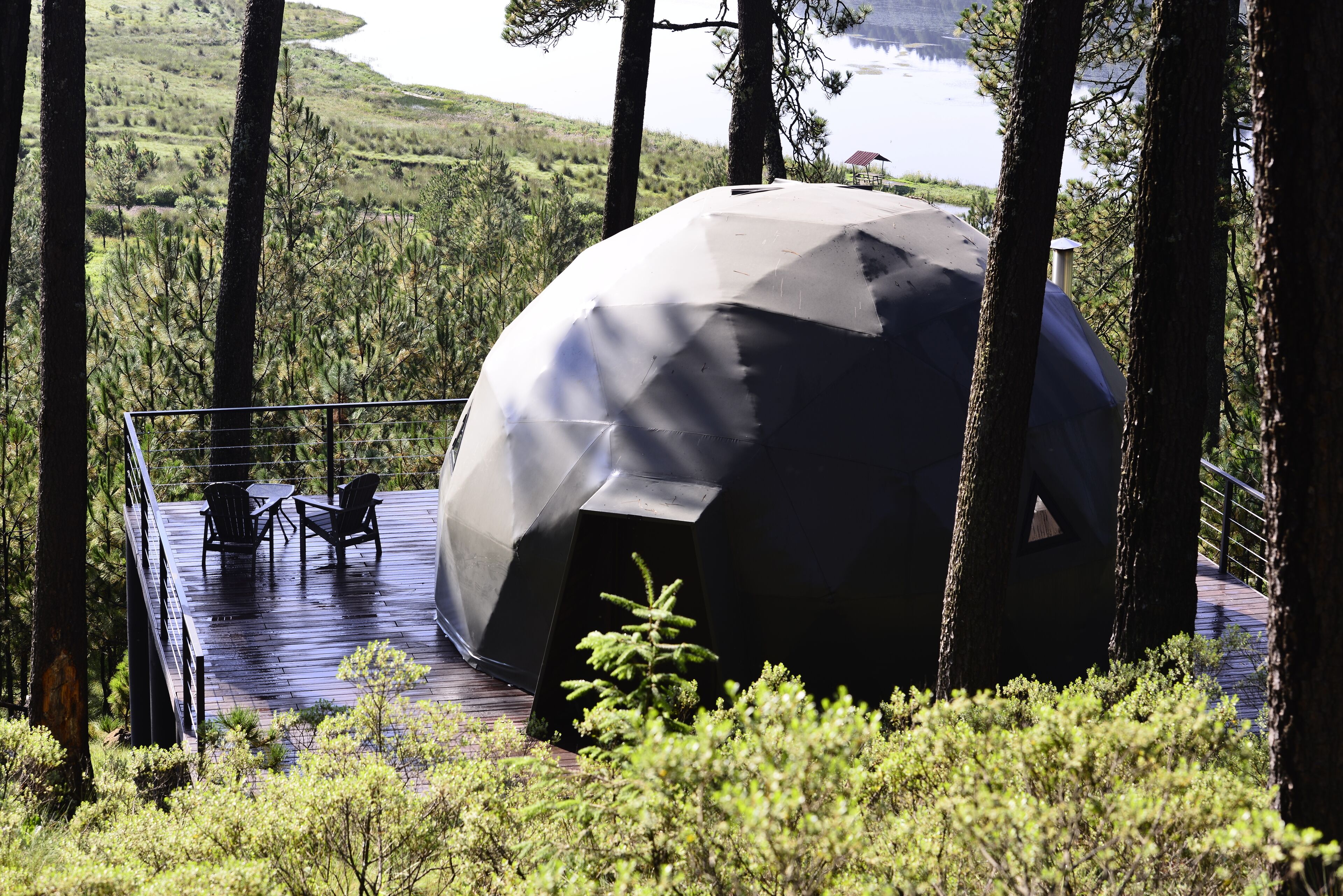 A geodesic dome structure with a deck and chairs, surrounded by trees and a view of a lake.