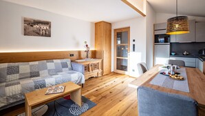 A modern living room with a wooden sofa, a dining table with chairs, a kitchen area, and a large window. - Apartment 'Hubertus - Wasserfallspitz' with Mountain View, Private Terrace and Wi-Fi (Sand in Taufers)