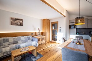 A modern living room with a wooden sofa, a dining table with chairs, a kitchen area, and a large window.