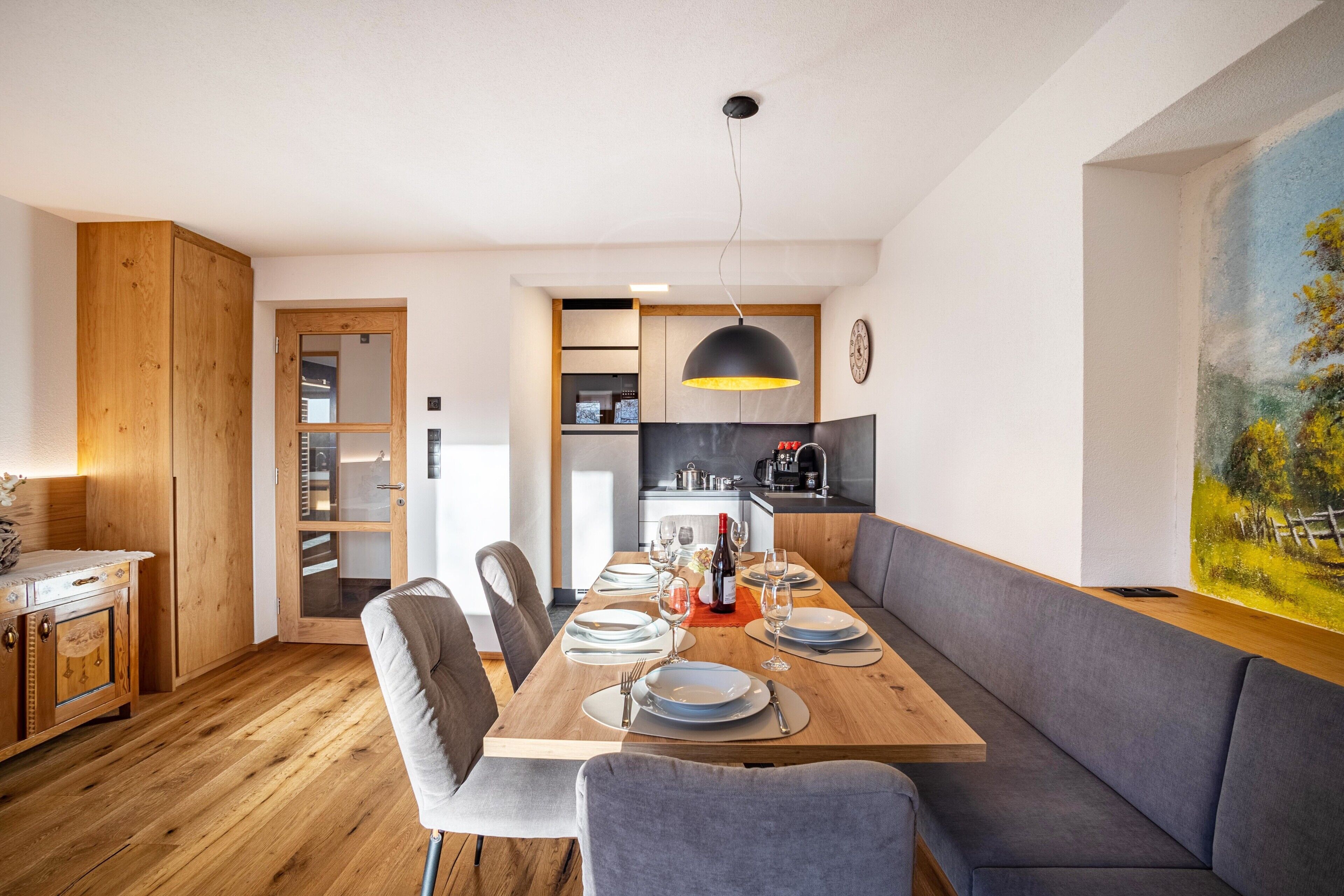 Un'area pranzo moderna con un tavolo in legno apparecchiato per quattro persone, un divano grigio e una cucina sullo sfondo.