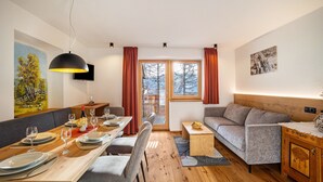 Een moderne woonkamer met een eethoek, houten vloeren, een bank, een aan de muur bevestigde televisie en een groot raam met gordijnen.