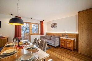 A dining area with a wooden table set for four, a sofa, a cabinet, and a large window with curtains.