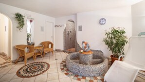 Ein Zimmer mit Korbstühlen, einer Mosaikfontäne, einer Topfpflanze und einer Wanduhr.
