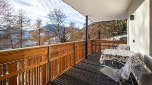 Ein Balkon mit Holzgeländer, einem Tisch mit blumigem Tischläufer und einem Sessel mit Felldecke.