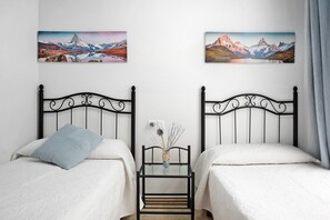Une chambre avec deux lits, une petite table sur laquelle se trouve un vase, et deux tableaux accrochés au mur.