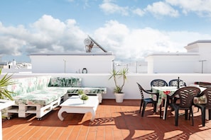 Eine Dachterrasse mit einer weißen Couch, einem Esstisch mit Stühlen, Topfpflanzen und einer Satellitenschüssel.