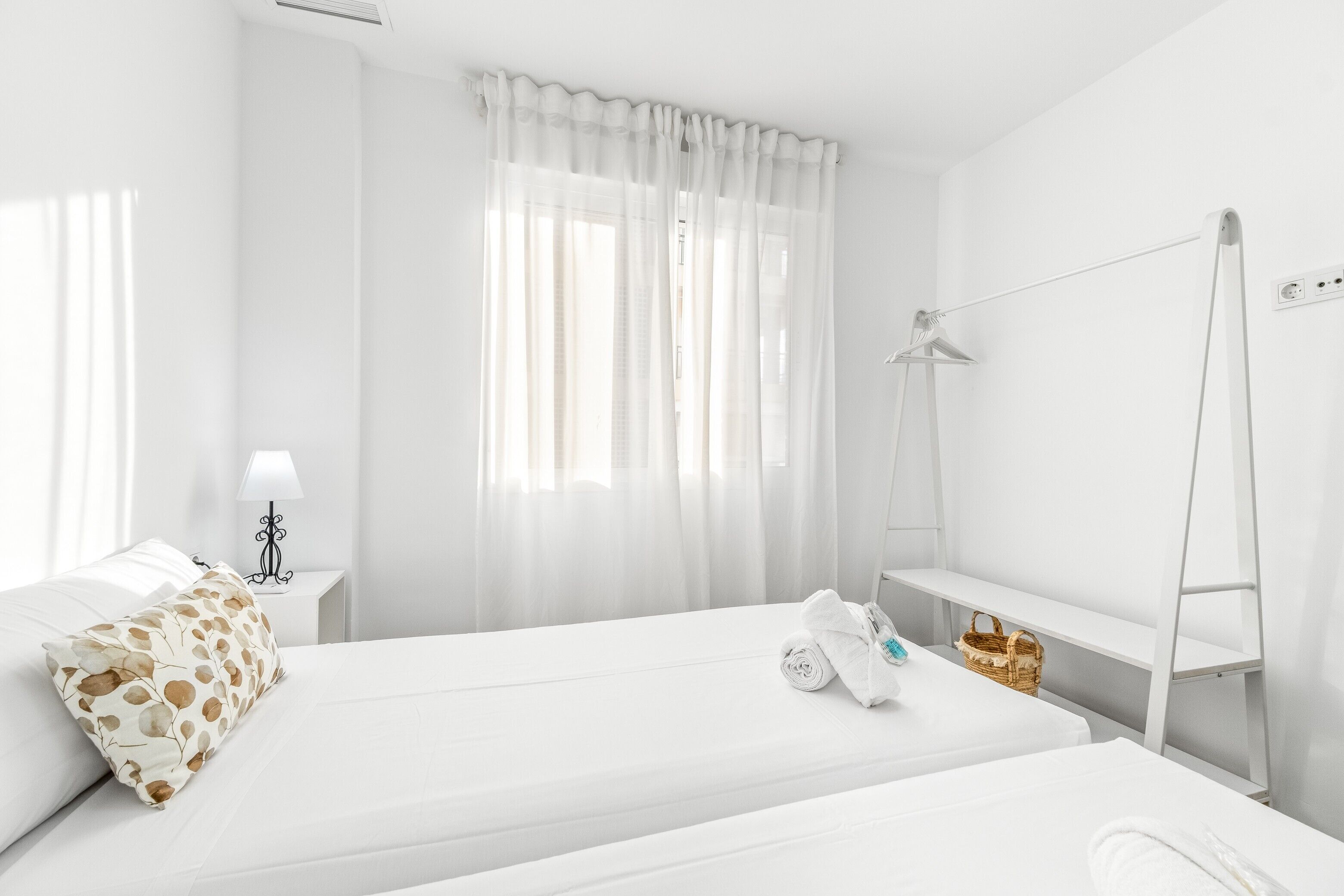 A hotel room with a bed, a nightstand with a lamp, a window with sheer curtains, and a wall-mounted shelf with a basket.