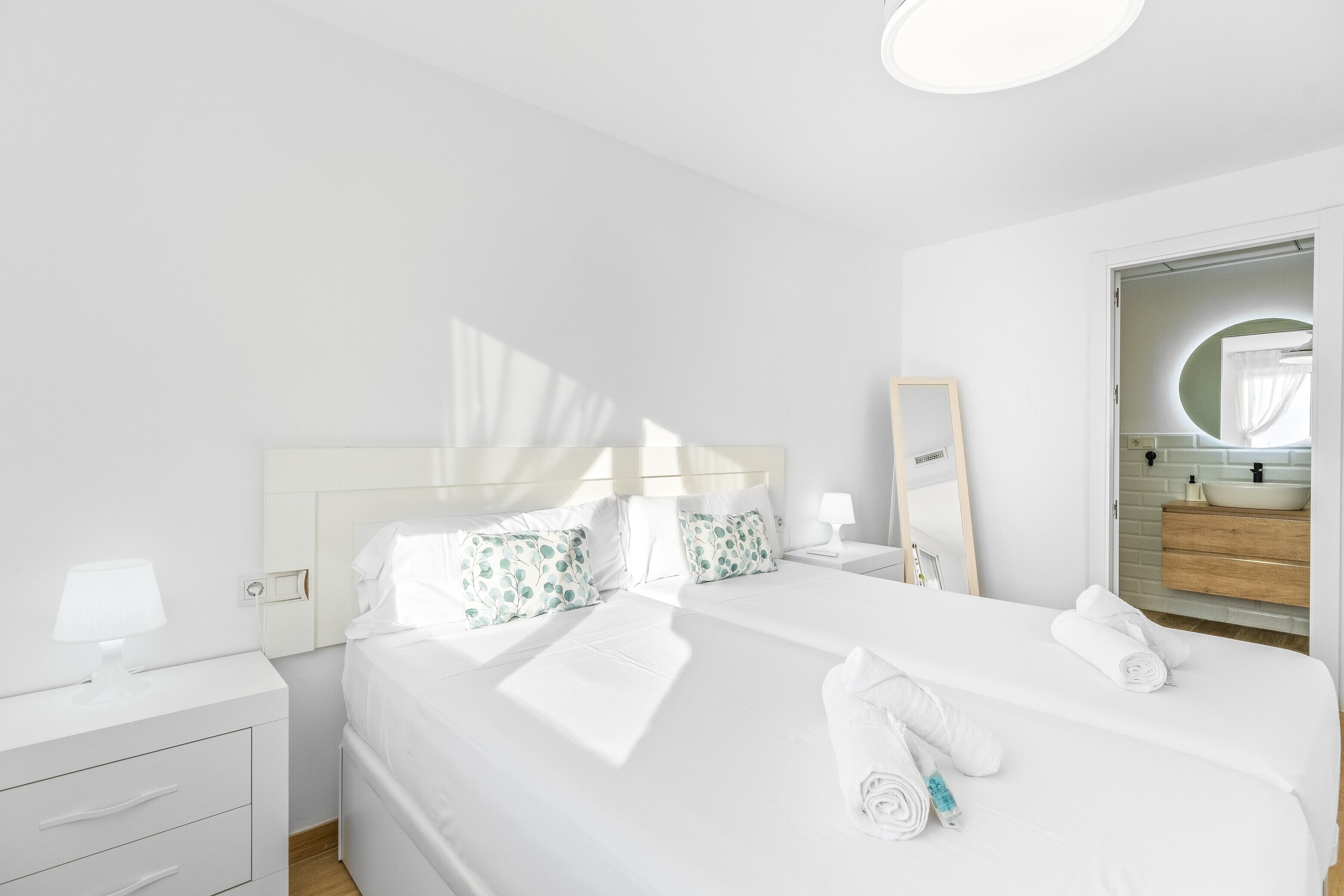 Une chambre d’hôtel avec un lit, une table de chevet, une commode, un miroir et une salle de bain visible à travers une porte ouverte.