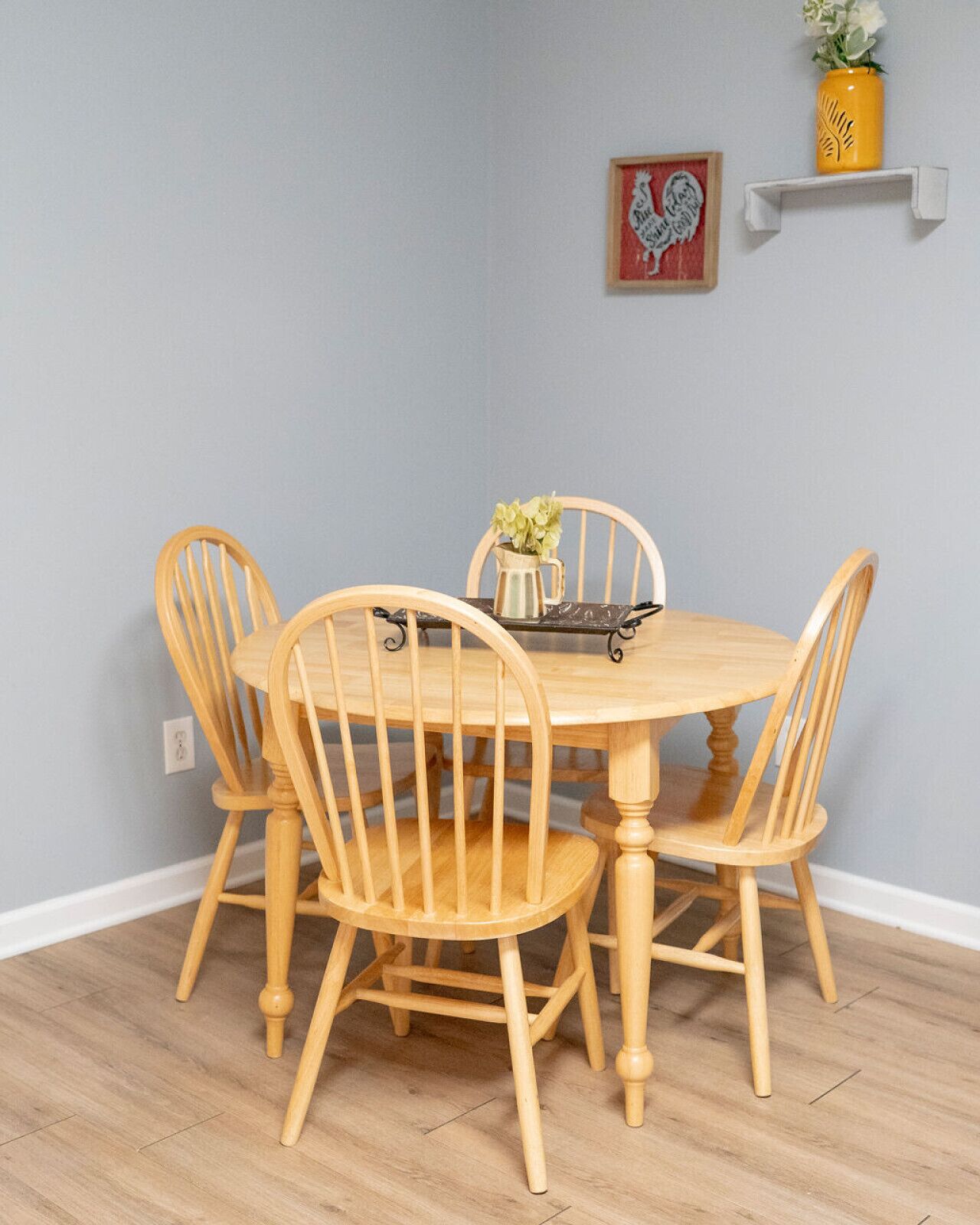 A dining table with four wooden chairs, a pitcher of flowers, and a framed picture on the wall.