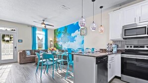 A modern kitchen with white cabinets, a built-in microwave, and a dishwasher. There is a dining area with blue chairs and a table, and a large window with curtains. - Beach house, 1.5 blocks from beach, 3bd, 3 bath (gulfport)