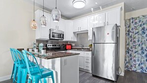 A modern kitchen with white cabinets, a stainless steel refrigerator, a microwave, and a stove. There are turquoise chairs around a small island. - Beach house, 1.5 blocks from beach, 3bd, 3 bath (gulfport)