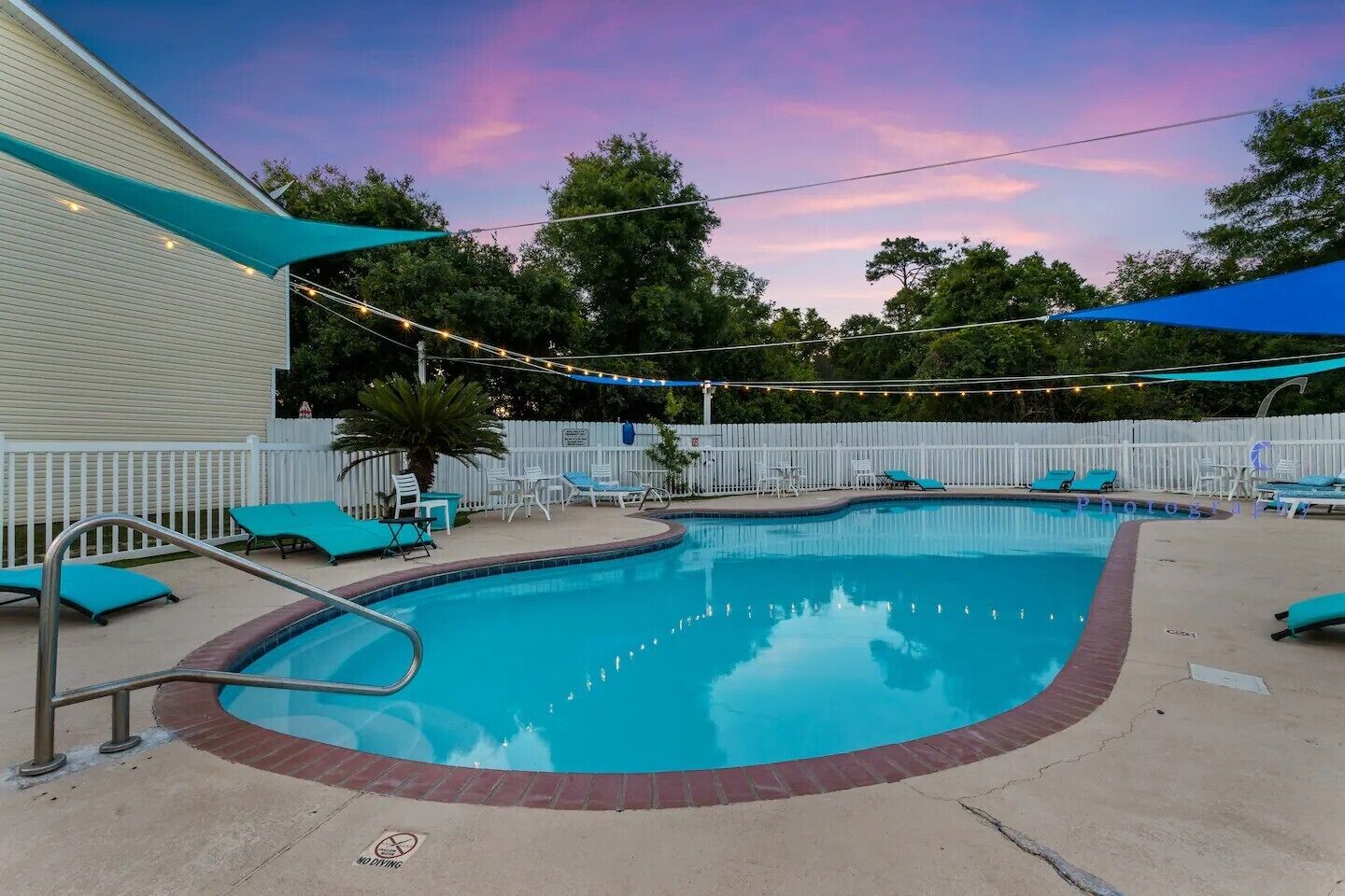 A swimming pool with a brick edge, surrounded by lounge chairs and a white fence.