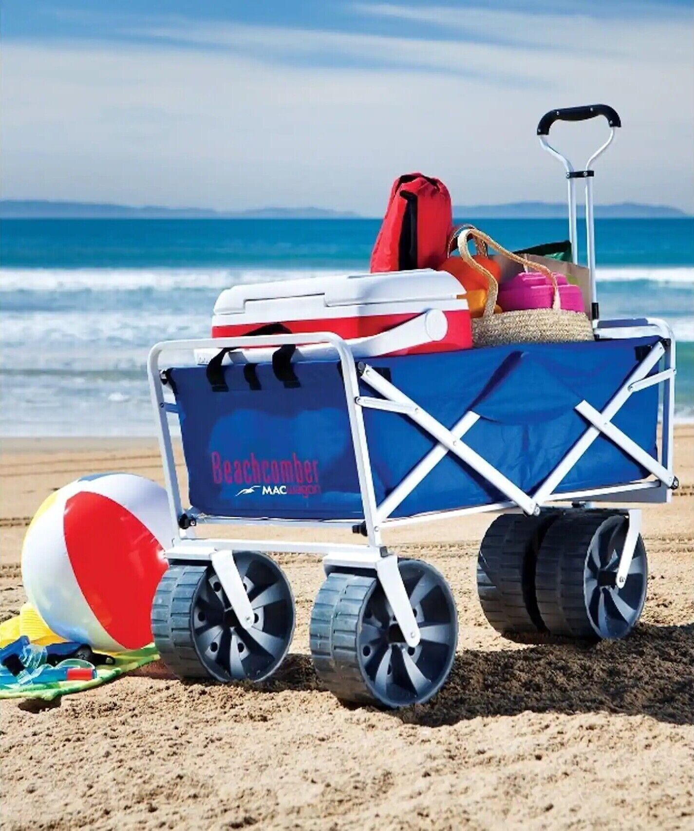 Een strandkar met een koelbox, een rode tas en een strandparasol op een zandstrand, met de oceaan op de achtergrond.