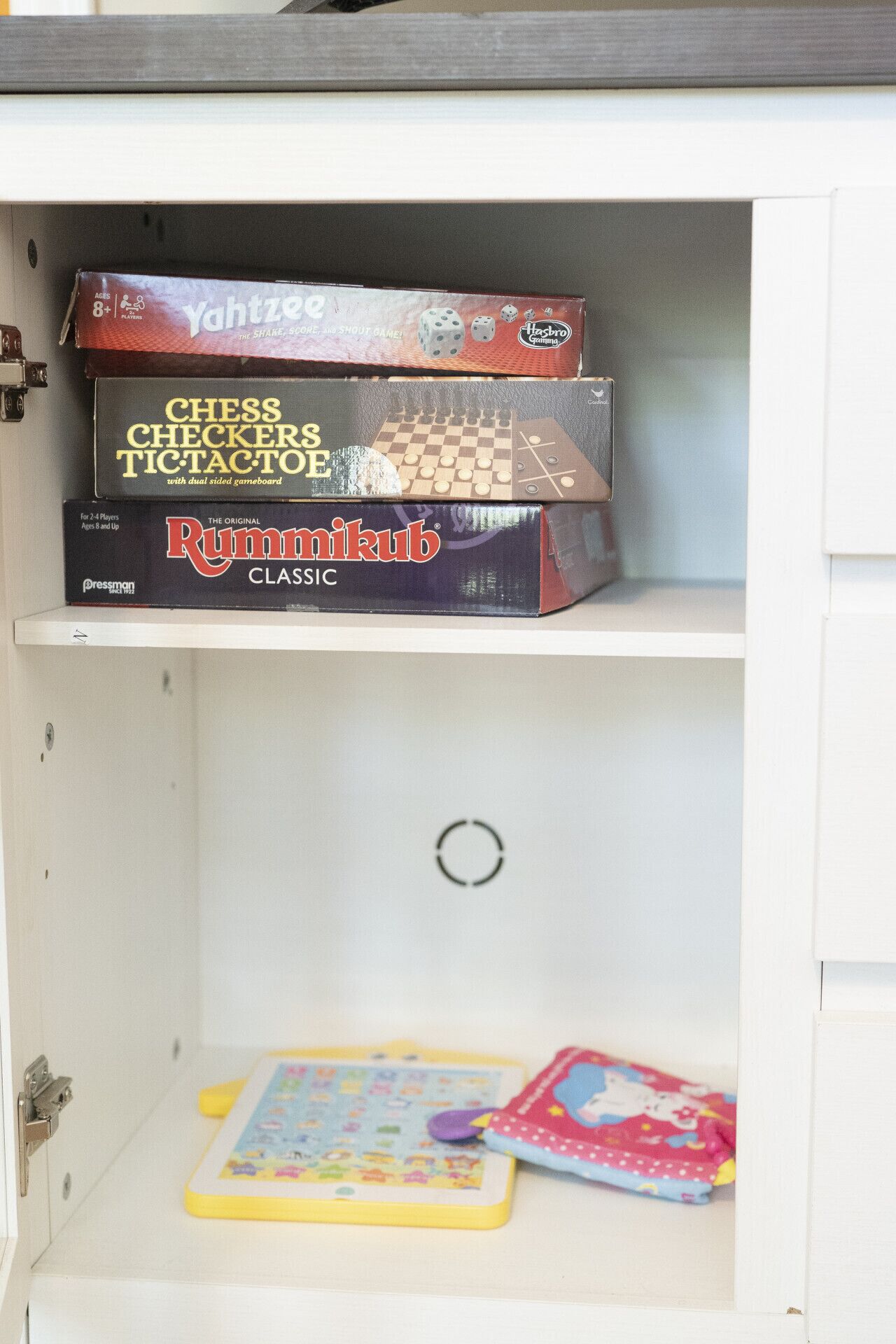 A cabinet with three board games: 'Yahizee,' 'Chess Checkers Tic Tac Toe,' and 'Rummikub Classic.'