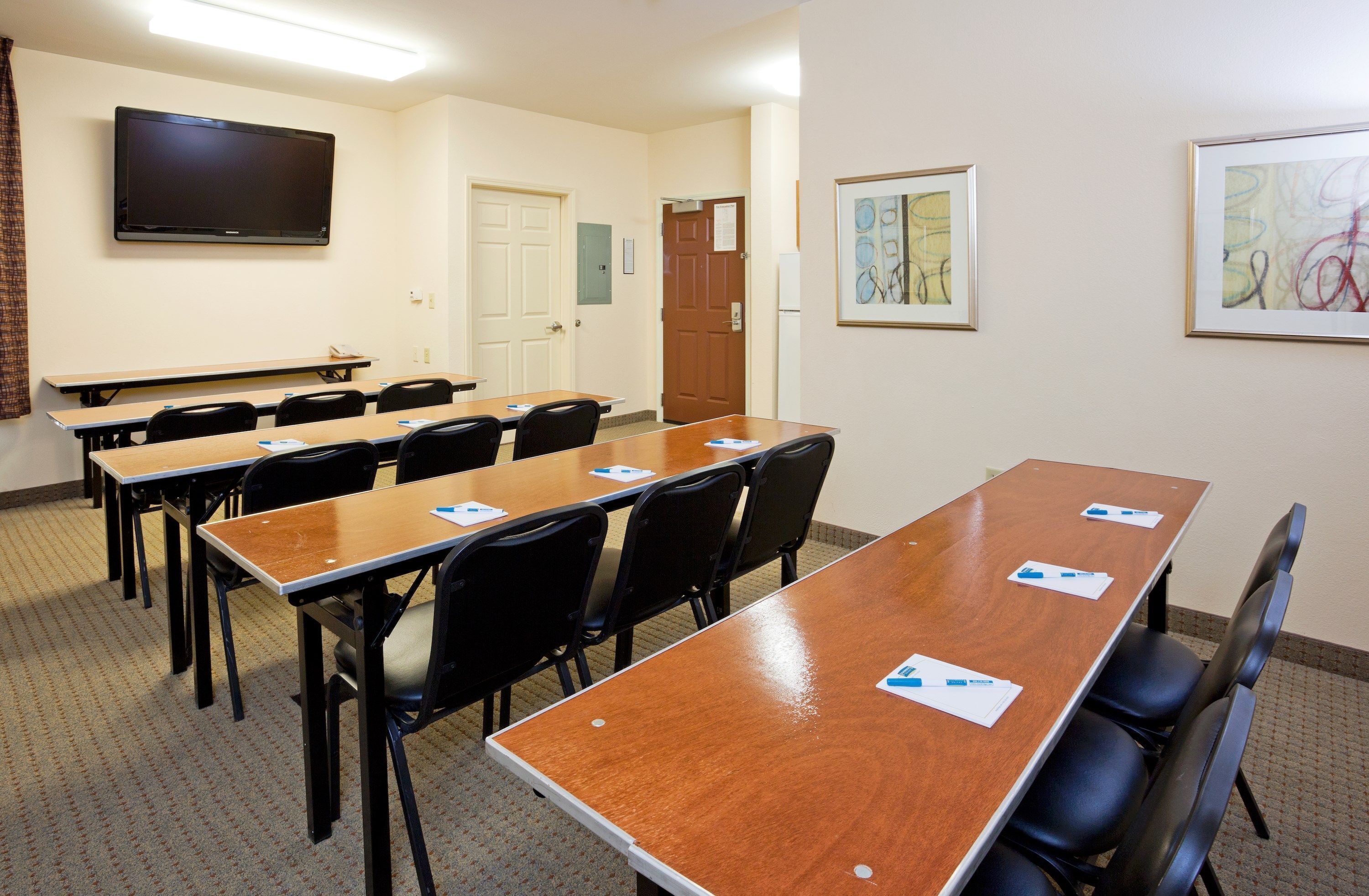 A conference room with long tables, black chairs, a flat-screen TV, framed artwork, and a door.