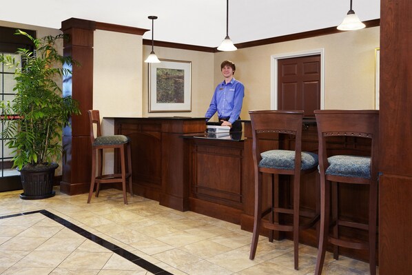 Zona de recepción con mostrador de madera, dos taburetes de bar, una planta en maceta y un cuadro en la pared.