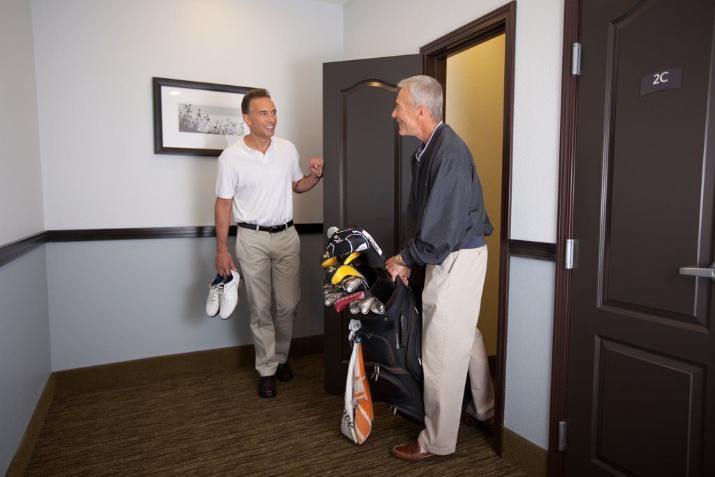 Two men in a hallway, one holding a golf bag and the other holding shoes.