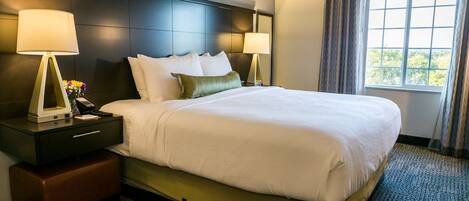 Ein Hotelzimmer mit einem großen Bett, zwei Nachttischen mit Lampen, einer kleinen Bank und einem Fenster mit Vorhängen.