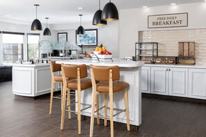 Una cocina moderna con una isla central, taburetes de bar, iluminación colgante y un letrero que anuncia desayuno diario gratuito.