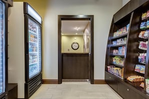 Ein Flur mit einem Kühlschrank voller Getränke, einer Wanduhr und Regalen mit Snacks und Pflegeprodukten.