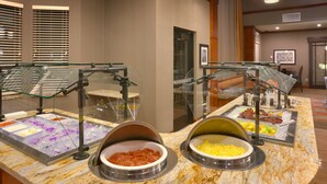 Uma área de buffet com diversas opções de comida, incluindo frutas, queijos e carnes, em um ambiente bem iluminado com bancada de mármore.
