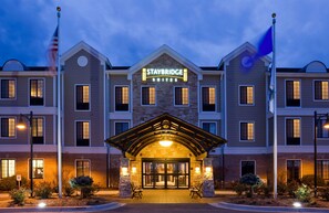 A Staybridge Suites hotel with a brick facade, a central entrance with a covered porch, and a flagpole with an American flag. - Accessible 2-BR Suite w/ Tub & 2 Bathrooms (Oconomowoc)