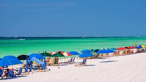 Uma praia com fileiras de espreguiçadeiras e guarda-sóis, céu azul e oceano ao fundo.