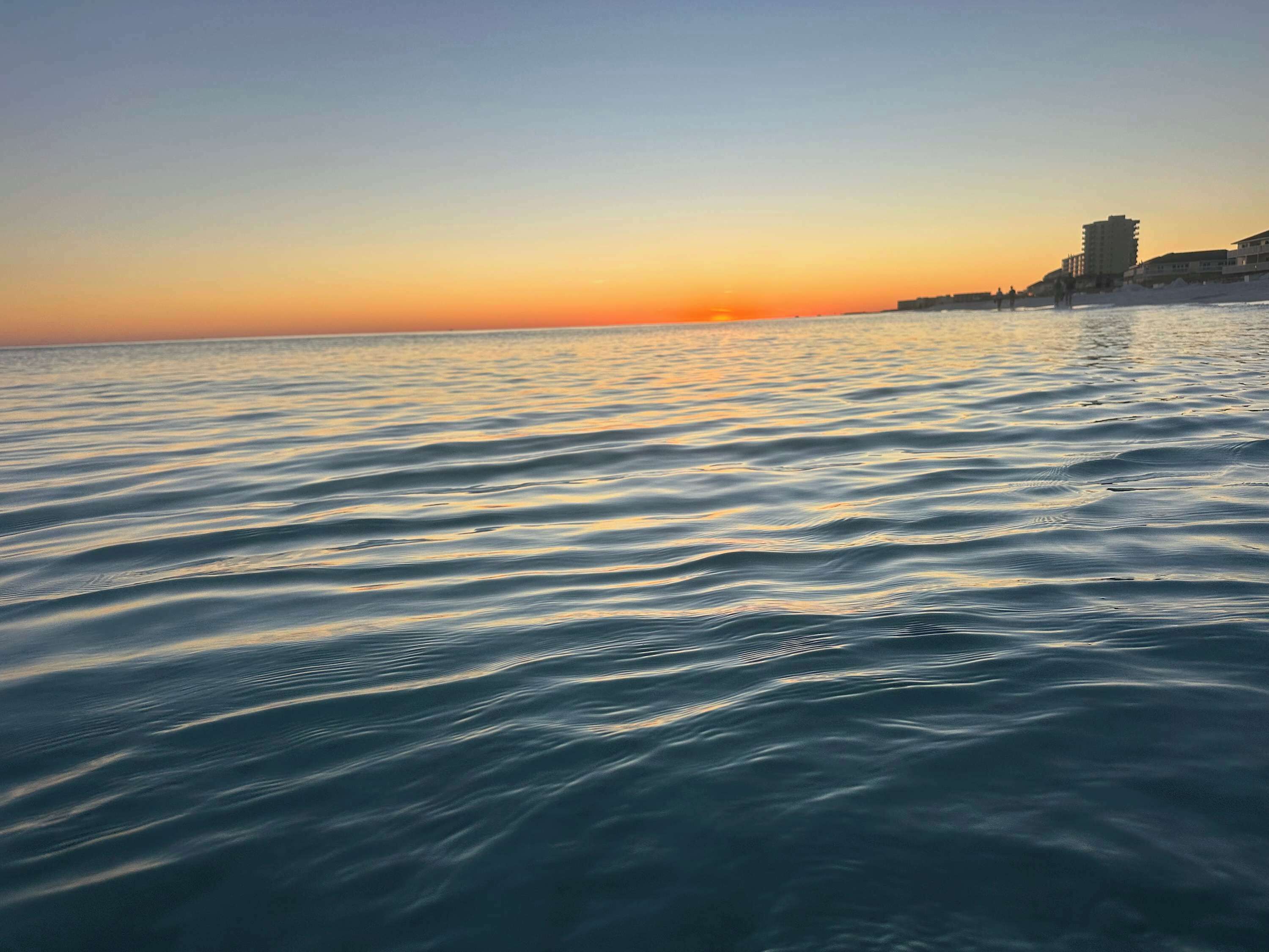 A calm sea at sunset with buildings in the background.
