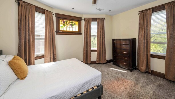 Un dormitorio con cama, cómoda, ventilador de techo y ventanas con cortinas.