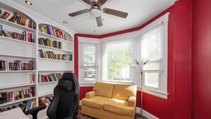 Una habitación con pared roja, molduras blancas, un sofá color beige, una silla de oficina negra y una estantería llena de libros.