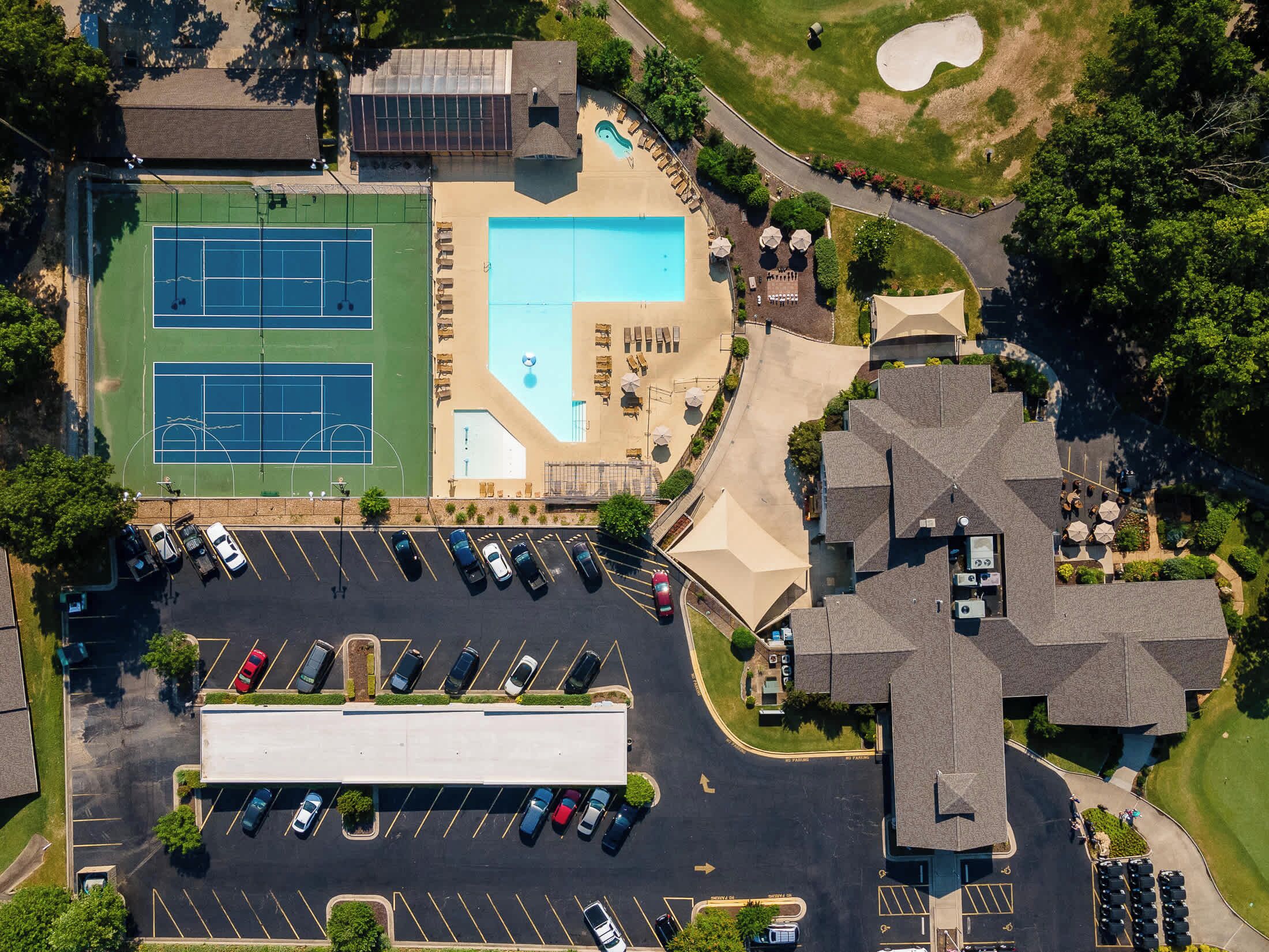 An aerial view of a property with a tennis court, a swimming pool, a parking lot, and a large building with a cross-shaped roof.