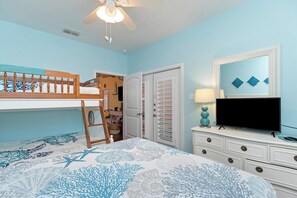 Quarto com cama beliche, escrivaninha, cômoda, televisão e ventilador de teto.