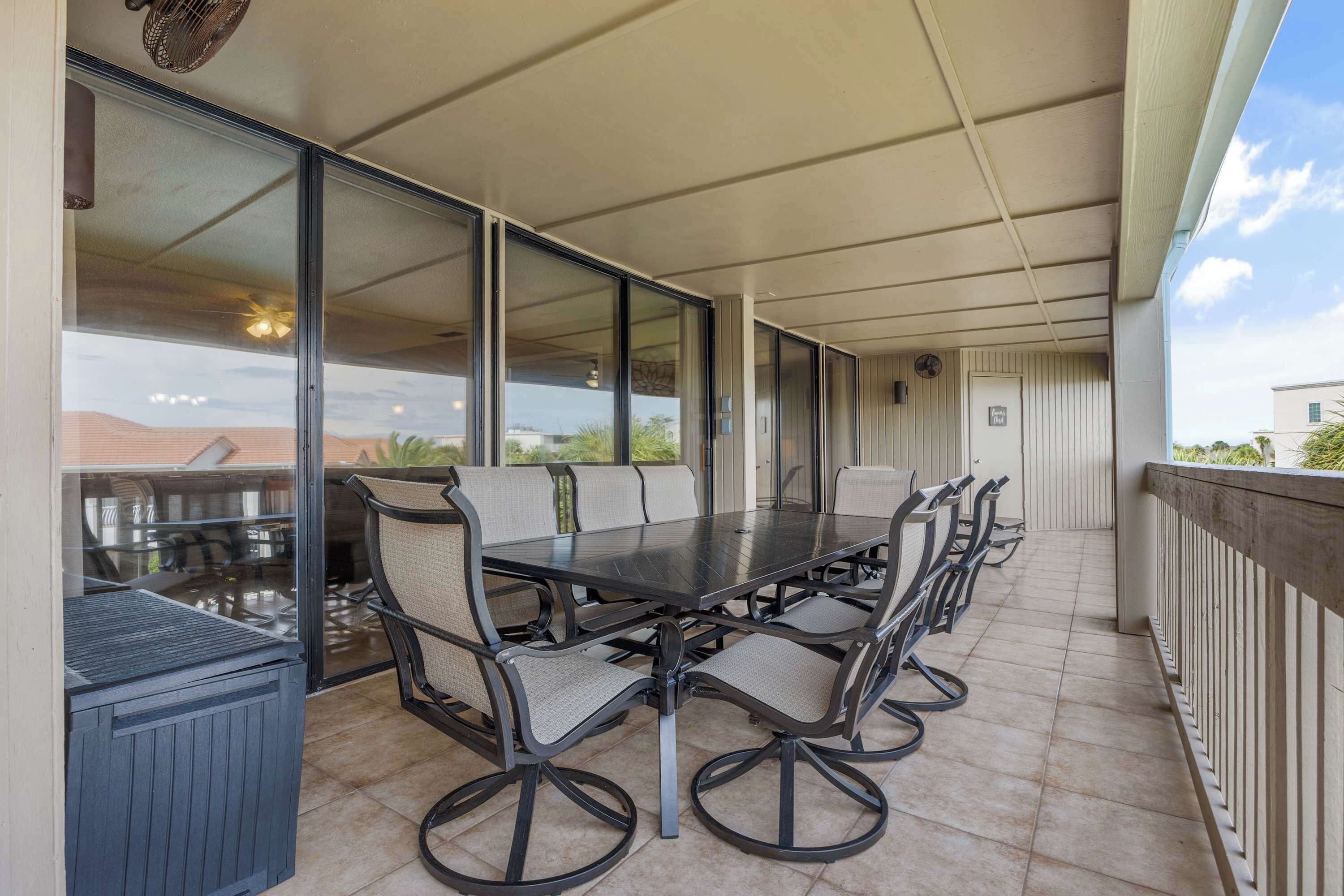 Uma área de refeições ao ar livre coberta, com uma mesa comprida e várias cadeiras, janelas amplas e vista para a paisagem.
